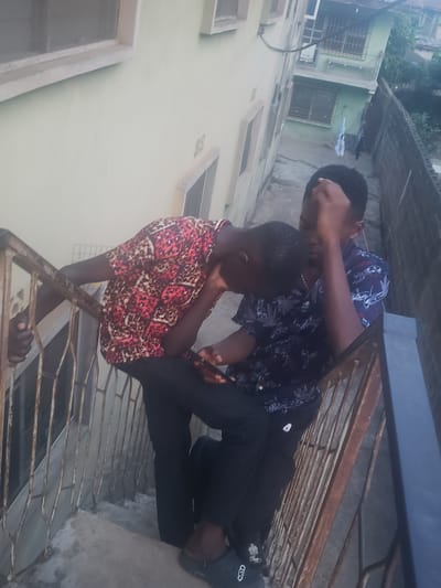 Two young men are standing on a concrete surface, likely a balcony or rooftop, in what appears to be a residential area. One man, wearing a red patterned short-sleeved shirt and black pants, is leaning slightly forward. The other man, dressed in a dark, floral-patterned shirt and black pants, is standing behind him. They are next to a metal railing, which has some rust. In the background, a building with light-colored walls and several windows is visible. Some foliage can also be seen in the distance. The location is presumed to be Alalubosa, Nigeria. The lighting suggests it is daytime.