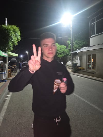 A young man stands on a street in Pripek, Bulgaria at night. He wears a black hooded sweatshirt and black pants. He holds an ice cream cone in his left hand and makes a "peace" sign with his right. A street lamp illuminates him and the surrounding area. Behind him is a building with illuminated windows and some greenery. A street runs beside him. In the background, there is what appears to be a street market or outdoor event with tables and people.