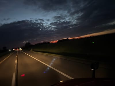 The image shows a view from a vehicle driving on a highway at dusk. The sky is filled with dark, dramatic clouds, with a sliver of orange and red visible on the horizon, suggesting the last moments of sunset. The road ahead is illuminated by the vehicle's headlights, with white lane markings clearly visible. In the distance, the lights of other vehicles and roadside structures can be seen. The right side of the image shows a dark, grassy embankment. There are no people visible in the image. The scene appears to be a typical highway landscape during twilight hours.