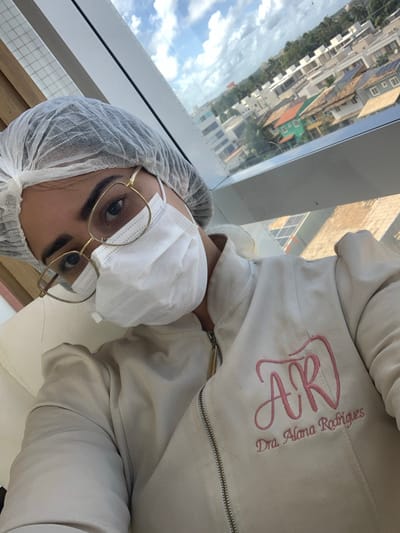 The image presents a close-up, elevated view of a female individual in an indoor setting within Lauro de Freitas, Brazil. The subject, likely a medical professional, wears a white disposable bouffant cap, a white medical-grade face mask, and gold-framed eyeglasses. She is dressed in a light beige zippered jacket, embroidered on the left chest with pink stylized initials "AR" above the text "Dra. Alana Rodrigues." The individual is positioned in a light-colored chair, looking directly towards the camera. Behind her, a large window provides an elevated perspective of an urban landscape under a partly cloudy sky. The visible exterior environment consists of a densely populated residential area with various low-rise and multi-story buildings, featuring diverse roof colors including red, green, and grey, some equipped with solar panels. Green foliage and trees are interspersed throughout the urban fabric. The scene depicts a professional environment with a view consistent with a high-rise building or clinic in a developed area of Lauro de Freitas.