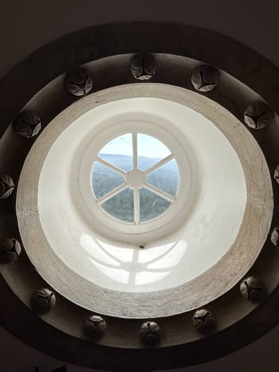 A low-angle, indoor shot showcases an ornate, circular window, presumably in a historical structure in Sintra, Portugal. The window itself is round with a white frame divided into six panes. Through the window, a landscape of green, forested hills and a sliver of a pale blue sky are visible. The window is set within a deeper, concentric ring of white plaster or stone, creating a narrow recess. The outermost ring, also circular, is made of a rougher, greyish material and adorned with a series of decorative elements resembling carved ornaments or emblems spaced evenly around its circumference. Light casts shadows within the recess of the window, creating a play of light and dark on the plaster or stone. The overall impression is one of age, architectural detail, and a connection to the natural world outside.
