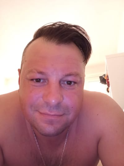 This is a close-up, selfie-style portrait of a man with a fair complexion and dark hair styled to the side. He is looking directly at the camera with a slight smile. The background is a plain, light-colored wall, suggesting an indoor setting. Some objects are visible to the right, including what appears to be a white cabinet or shelf with some colorful items on it. The lighting is warm, and the overall mood is casual and relaxed. The man is wearing a silver chain necklace. The image is likely taken indoors during the day, as indicated by the lighting. No text is visible in the image.