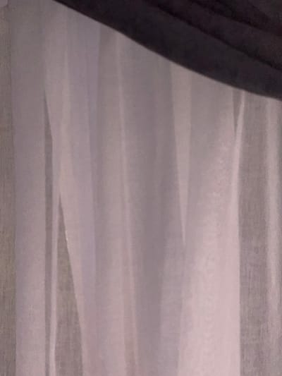 The image shows a close-up of draped fabric, likely curtains or a soft furnishing. The fabric is a light, translucent material, possibly sheer or semi-sheer, in shades of white and light pink, with vertical folds and pleats that create subtle shadows and highlights. A darker, opaque material, possibly black or dark gray, is visible along the top edge of the frame, casting a shadow onto the lighter fabric below. The texture of the fabric appears soft and slightly textured, with some visible grain or speckling, possibly due to the lighting or the material itself. There are no people, animals, or specific objects clearly identifiable beyond the fabric. The background is indistinct, suggesting a shallow depth of field or the nature of the material obscuring whatever is behind it. The scene is static and appears to be indoors. The provided location information "Doma 1096 Km, Russia" is not visually derivable from the image itself.