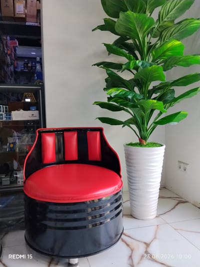 A red and black barrel-shaped chair with red padded backrests sits in front of a tall potted plant with large green leaves. The plant is in a white textured pot. To the left of the chair, a metal shelf unit filled with various items, including boxes and bottles, is visible. The floor is tiled with a marbled pattern. An electrical outlet is present on the white wall behind the plant. The image includes a timestamp at the bottom right corner reading "23.06.2026 10:00" and "REDMI 15" at the bottom left. The overall scene appears to be an indoor setting, possibly a storage area or a workshop in Yovon, Tajikistan.