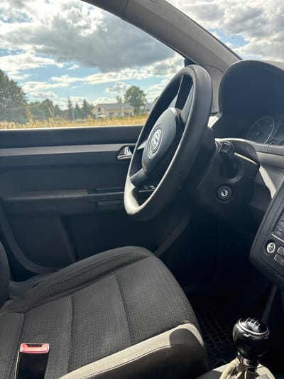 The image shows the interior of a car, a Volkswagen, from the passenger seat, looking towards the driver's side. The dashboard and steering wheel are visible, with the gear shift in the center console. Outside the car window, a rural landscape is visible with trees, a field of dry grass, and a house in the distance under a partly cloudy sky. The location is identified as Mikorzyn, Poland, implying this is a view from a vehicle parked or driving in this area. The overall scene is one of a sunny day in a rural setting, viewed from inside a car.