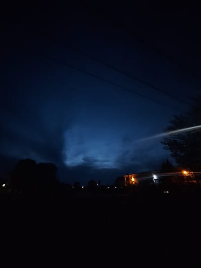 The image depicts a night scene in Dolni Chiflik, Bulgaria. The view is from a low angle, likely street level, looking up at the sky. The sky is a dark blue with areas of lighter, cloudy patches. Power lines are visible in the upper left portion of the frame, extending diagonally across the scene. Along the right edge of the image, there is a line of trees and buildings, obscured by darkness, with some lit windows and streetlights appearing as small, bright orange and white dots.