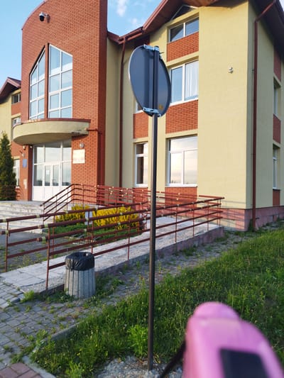 The image shows the exterior of a building in Novyi Rozdil, Ukraine. The building has a light yellow facade with red brick accents and a brown roof. There are several windows with white frames visible. In front of the building is a paved walkway with a ramp and handrails. A concrete trash can with a black plastic bag is located on the walkway. A black pole with a circular sign is in the foreground, partially obscuring a pink object. The area is landscaped with green grass and some yellow flowers. The sky is blue with some clouds.