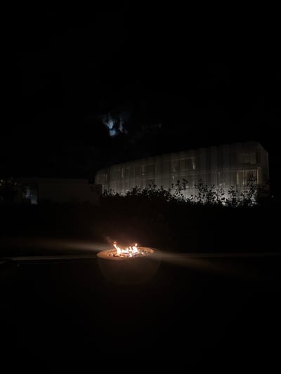 At night, a glowing fire pit is on the right. A tall building with linear, grid-like windows is on the left, partially obscured by dark foliage. A hazy, illuminated moon is visible behind dark clouds in the background. The scene is dark, with the fire pit and building casting the only significant light.