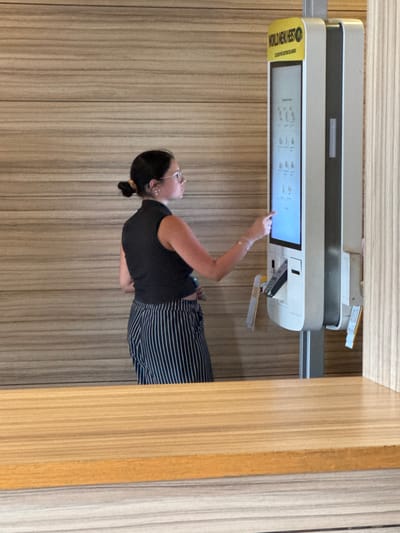 The image shows a woman interacting with a touchscreen kiosk. The woman is wearing glasses, a black tank top, and striped pants. She is standing and reaching out to the screen with her right hand. The kiosk is a light gray color and has a screen displaying various icons and text. A yellow sticker with black text is visible on the left side of the kiosk. The background appears to be a wall with a wood-like texture. The overall lighting suggests it is daytime. The scene is likely in a public indoor space, possibly a restaurant or retail establishment. The woman appears focused on the task at hand. The text on the yellow sticker reads "WORLD MENU" and "OCCHIO ALLE OFFERTE."