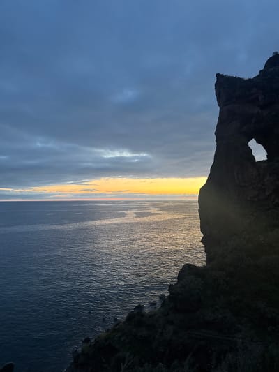 Here is a description of the image:
The image shows a view of the ocean and sky from a rocky cliff in Ponta do Sol, Portugal. The foreground is dominated by the dark, jagged silhouette of the cliff face, with a small opening visible through the rock. Beyond the cliff, the ocean stretches out, its surface reflecting a vibrant sunrise or sunset, with a band of yellow and orange light. The sky is a mix of dark blues and grays. The image is rotated 90 degrees counter-clockwise.