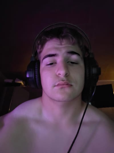 A young Caucasian male with fair skin, dark curly hair, and dark eyebrows is looking directly at the camera. He is wearing black over-ear headphones. The image is lit with purple and pink lighting. The background is dark and out of focus, with some objects visible on the left. The male appears to be shirtless, with his shoulders and upper chest visible. The scene is a close-up, head-and-shoulders shot. The location is not discernible.