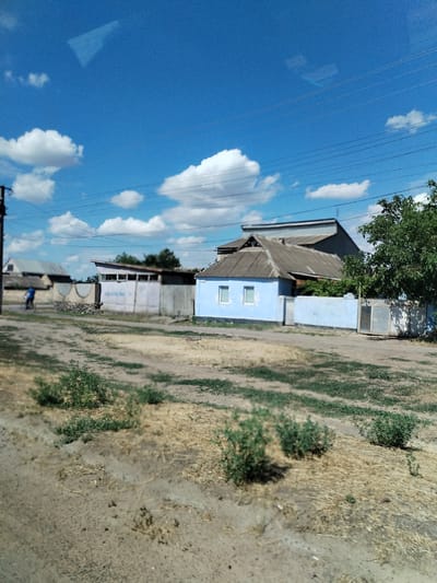 The image depicts a street scene in Nova Odesa, Ukraine, under a bright blue sky with scattered white clouds. The foreground shows a dirt road with patches of dry grass and some green bushes. To the left, a utility pole stands next to a fence. A person on a bicycle is visible in the mid-ground, riding along the road. Behind the fence, a light blue building with two white-framed windows is prominent. Adjacent to it is a longer, pale purple building with a sloped roof, partially obscured by trees. Further in the background, other structures and vegetation can be seen. Power lines stretch across the sky. The overall scene suggests a rural or semi-rural residential area.
