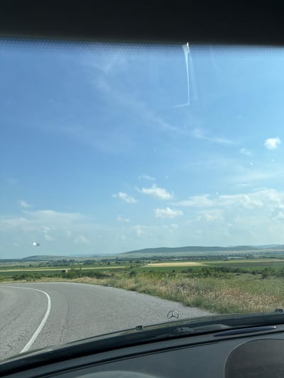 The image, captured from the interior of a vehicle, presents a wide-angle view of a bright, sunny daytime scene in Kovachite, Bulgaria.

The upper part of the frame is dominated by a clear blue sky, interspersed with scattered white cumulus clouds, indicating fair weather. The sun is high, illuminating the landscape brightly.

The lower portion of the image displays a rural setting. A paved road, with a distinct solid white line marking its edge, is visible on the right side, curving gently into the distance. This road is flanked by expansive green fields and meadows, with varying shades of green suggesting different types of vegetation or agricultural land. In the far distance, a hazy horizon line reveals the gentle contours of mountains or hills, blending softly with the sky.

Reflections are visible on the car's windshield. On the left side, a dark area, likely part of the car's interior trim or dashboard, is reflected along with a distinct white, rectangular object that has a small, flag-like protrusion. On the far right, partially visible on the car's interior structure, is the iconic shiny three-pointed star emblem, characteristic of a Mercedes-Benz logo, identifying the vehicle's make.

No people are visible in the scene. The image evokes a sense of peaceful travel through scenic countryside during good weather conditions.