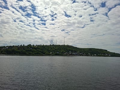 A wide, dark grey river fills the foreground of the image, with small ripples reflecting the light. In the background, a green, tree-covered hill slopes down towards the water. Several houses and buildings are visible scattered across the hillside. Two tall communication towers stand prominently on the crest of the hill, one to the left and a taller one to the right. The sky above is a bright blue, filled with a dense scattering of white, fluffy clouds. The image appears to be taken from a boat on the river, looking towards the town of Verhnij Uslon, Russia.