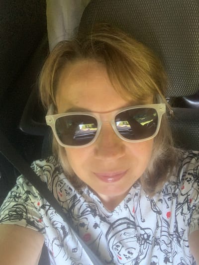 In a medium shot, a woman with blonde hair is captured inside a vehicle. She is wearing sunglasses and a white t-shirt with a black and white line drawing of faces. The sunglasses reflect a road with trees and cars, suggesting the vehicle is in motion. The background is dark, possibly the interior of the car, with a textured seat visible. The image is likely taken in Слабада-Кучынскі сельскі Савет, Belarus, given the user's prompt.