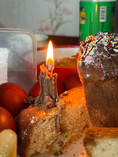 A close-up indoor shot shows food, likely bread or cake, in front of a lit candle with dripping wax, resting on what appears to be dyed eggs. The cake has sprinkles and chocolate and is partially cut, revealing the crumb. The candle is almost entirely melted, with the wax running down the cake. The background is blurry, with a container and part of a can visible. No people are visible in the frame. The location is not possible to determine from the image itself, though Пожарское сельское поселение is a potential location based on the request.