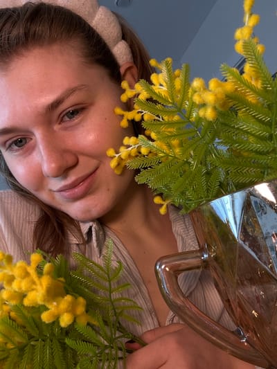 A woman's face, angled, is visible among green and yellow foliage. Her complexion is fair, with clear skin and visible eyebrows and eyelashes. She wears a light-colored headband. Her lips are slightly parted in a smile. Above her, a transparent glass object with a metallic handle reflects the surrounding environment. She is holding the flowers in her right hand. The scene appears to be an intimate, close-up perspective.