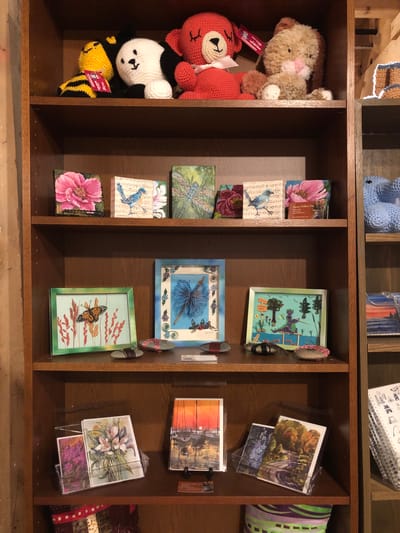 The image displays a wooden bookshelf filled with various decorative items and artwork.

On the top shelf, there are three crocheted stuffed animals: a yellow and black bee, a white and black panda, and a red bear with a pink bow. To the right of the red bear is a brown and cream colored stuffed bunny.

The second shelf from the top features several small square artworks. From left to right, there is a painting of a pink flower, a painting of a blue bird on sheet music, and a painting of a dragonfly.

The third shelf from the top showcases framed artwork. On the left is a framed picture of a monarch butterfly on a stalk of wheat. In the center is a blue framed piece with what appears to be a dragonfly made of intricate paper or thread work. To the right is a framed picture of a small green dragon sitting on a pier, fishing in a blue pond, with trees and fish visible. There are also several decorated stones on this shelf.

The bottom shelf holds several greeting cards and notebooks. On the left, there are three greeting cards displayed in a clear plastic stand, featuring floral and landscape paintings. To the right of these cards is a notebook with a spiral binding and drawings on its cover. Further to the right, more greeting cards are visible, and at the very bottom, part of a striped cushion or fabric can be seen.

The bookshelf itself is made of dark wood. The background to the left of the bookshelf appears to be a textured wall, possibly plaster or concrete. The overall lighting suggests an indoor setting, likely a retail space or a home.

There are no people visible in the image. The items are arranged in an organized manner on the shelves. The location is identified as the city of Severn, Canada.