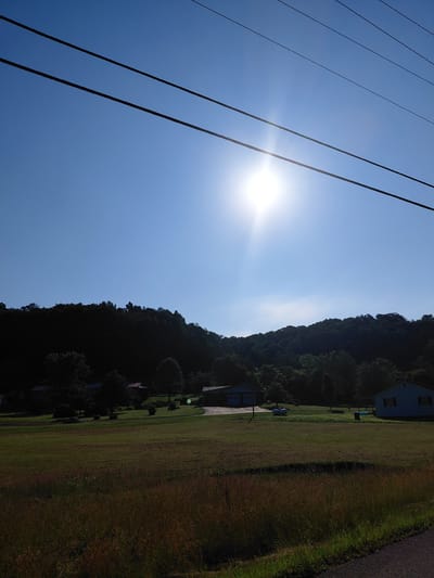 This is an eye-level, outdoor, full shot of a sun-drenched rural landscape. The sky is a clear, bright blue with a prominent sunburst effect in the upper center. Four dark power lines stretch horizontally across the top of the frame. In the middle ground, a gentle slope is covered in a lush green lawn dotted with trees and a few houses, including a light blue house on the right with dark shutters and a blue house with a garage door on the left. A light blue cylindrical object is visible on the lawn between the two houses. The foreground consists of a field of tall, dry grass with hints of green and brown. A paved road is visible on the far right edge of the frame. The background is a dense, dark green forest that covers the rolling hills. The image appears to be taken in Berea, United States, based on the provided context.