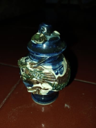 A small, lidded ceramic vessel is positioned centrally on a reddish-brown tiled surface. The vessel, approximately 12 cm in height, features a predominantly dark blue glaze with sections of white, dark brown, and dark green. It is adorned with intricate relief ornamentation, depicting a stylized dragon or mythical creature wrapping around its main body, exhibiting textured scales and defined limbs. The white glazed areas show visible crackling (crazing). The vessel has a somewhat globular body, a distinct flared base, and a cap-like lid with a small, rounded finial. The tiled surface beneath the object displays visible grout lines and minor light-colored particulate matter. The background is largely dark and out of focus, indicating an interior setting with localized illumination. The scene is located in Longmiang, Laos.
