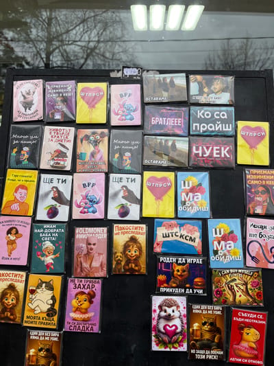 A close-up, eye-level shot captures a collection of approximately 50 to 60 rectangular magnets displayed vertically on a black, possibly metallic, board. The scene is likely located in a shop or stall in Rakitovo, Bulgaria. Each magnet features a distinct illustration or phrase, predominantly in Bulgarian Cyrillic, encased in clear plastic. Themes vary widely, including cartoon characters like Stitch, anthropomorphic animals (pigs, cats, hedgehogs, squirrels), stylized human figures, and various objects like balloons or Nutella jars. Many magnets contain humorous or colloquial phrases. A price tag is affixed to the upper right portion of the board, displaying "€ 2.00" and "лв./бр." (BGN/piece). The background, visible as a reflection on a glass surface behind the magnets, shows blurred outlines of bare tree branches against a light, possibly overcast, sky. Four rectangular white light fixtures are also clearly reflected at the top of the glass. No people are directly visible in the immediate foreground, but the merchandise is arranged for viewing and sale.