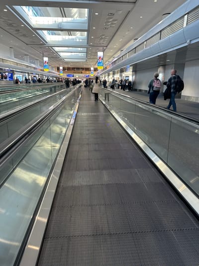 An interior shot captures a long moving walkway in an airport or similar public space. People are sparsely spread along the walkway, with a small cluster at the far end. Large windows are visible on one side. Signage hangs from the ceiling advertising gates. The walkway's surface is lined with horizontal grooves. The lighting is bright, and the overall atmosphere is sterile.