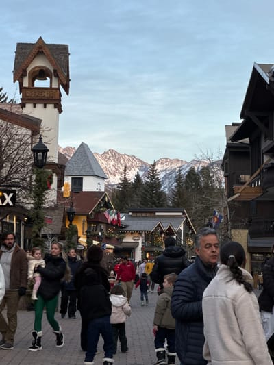 A scenic mountain town scene with numerous people walking along a brick-paved street. Buildings with unique architecture frame the sides, some adorned with Christmas decorations. Snow-capped mountains rise in the background under a cloudy sky. The perspective is rotated slightly, offering an interesting view of the bustling town amidst the natural landscape.