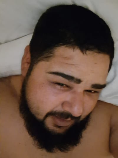 The image is a close-up, low-angle shot of a man lying in bed, seemingly just waking up. His head is tilted back, and his eyes are partially closed and appear tired. He has dark hair, thick eyebrows, and a full beard and mustache. The bedding is white and slightly wrinkled. The lighting is soft and appears to be from an overhead source. The scene is domestic and intimate, suggesting a private moment. The image does not provide any visual clues to its specific location in Thüngersheim, Germany, or any other city.