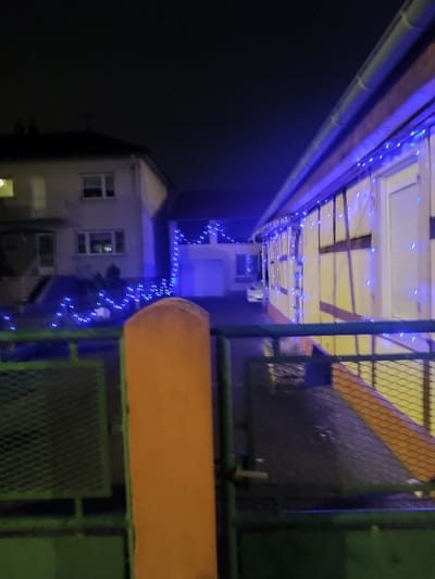 A dark night scene showcases two buildings illuminated with blue lights. The building on the left appears to be a two-story residential home. To the right, a building with half-timbered architecture is draped in strings of blue lights. A green fence with an orange post stands in the foreground. The wet pavement reflects the lights, contributing to the scene's overall brightness despite the darkness.