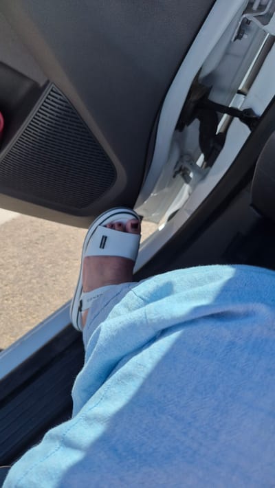 A first-person perspective shot shows a person's leg and foot inside a vehicle. The person is wearing light blue linen pants and a white sandal with a thick sole. The sandal has a single white strap across the toes and a black stripe along the edge of the sole. The foot is positioned on the car's door sill, with the car door ajar. The interior of the car is visible, including a dark gray door panel with a speaker grille. The exterior of the car is a bright white. The scene is likely in a parking area or on a street in Līvāni, Latvia, as indicated by the context.