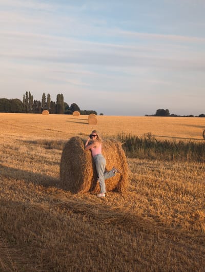 A young woman with blonde hair and sunglasses is posing next to a large, round bale of hay in a wheat field. She is wearing a pink tank top and ripped denim jeans. Her right leg is bent at the knee and extended backward, and she is leaning on the bale with her arms. She appears to be holding a camera to her face, as if taking a photo or being photographed. The field is golden brown, indicating it has been harvested, and several other hay bales are visible in the distance. The sky is a pale blue with wispy clouds, and the sun is casting long shadows across the field, suggesting it is late afternoon or early evening. The scene is set in Alvimare, France.