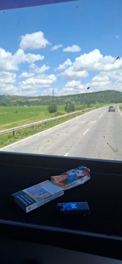 The image shows a close-up view of a car's interior, specifically the dashboard and windshield from the passenger side. Through the slightly dirty windshield, a sunny day scene unfolds with a clear blue sky filled with fluffy white clouds. A highway stretches into the distance, bordered by lush green fields and trees, suggesting a rural or semi-rural setting. The perspective is angled, with the road appearing to recede diagonally.

On the dashboard, a pack of cigarettes, some of which are visible through a plastic wrapper with orange and blue accents, sits next to a blue lighter. The cigarette pack has text on it, including what appears to be health warnings in a non-English language, possibly Cyrillic. A single cigarette pack is also visible on the right side of the frame. The overall impression is a snapshot of a road trip, perhaps in Bulgaria given the location context, during daylight hours under fair weather. The details of dirt on the windshield and the placement of the cigarettes suggest a casual and perhaps slightly cluttered environment within the vehicle.
