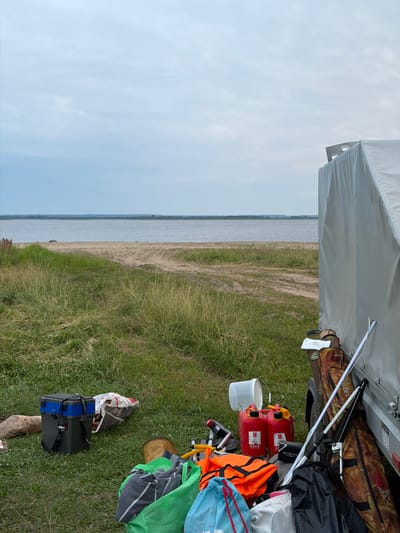 The image shows a collection of camping gear and supplies on a grassy area near a body of water. A large, light-colored tent is visible at the top of the frame. Scattered around on the ground are various items including two red jerry cans, a white bucket, an orange life vest, a green bag, a blue drawstring bag, a camouflage-patterned duffel bag, a black bag, and two other bags, one of which appears to be a cooler with blue straps. There's also a fishing net and what looks like fishing rods.
The scene is set outdoors, with a wide expanse of water and a sandy shore in the background. The sky is overcast, suggesting a cloudy day. The grass appears somewhat dry in places, indicating it might be a warm season. The location is identified as Красный Бор, Russia. No people are visible in the image, and the focus is on the gathered equipment, implying preparation for or the aftermath of an outdoor activity like camping, fishing, or boating.
Notable visible text includes "AQUATIC" and "EXTENTA" on some of the items, though the full context is unclear. The overall impression is one of readiness for an outdoor adventure by the water.