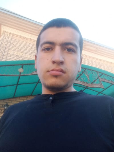 A frontal, waist-up portrait depicts a young adult male in the city of Kasan, Uzbekistan. The subject, centered in the frame, has short dark hair, dark eyes, and light stubble on his chin and upper lip. He wears a dark, possibly black, short-sleeved collared shirt with two snap buttons visible at the neck. His expression is neutral. The background features a light-colored brick wall with a geometric pattern, topped by a lighter-toned cornice. A green, semi-translucent canopy or awning with a dark grid-like support structure and ornate dark metalwork is positioned behind the subject's shoulders. A wall-mounted light fixture is partially visible on the left. The upper background shows a bright, clear sky.