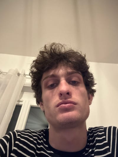 The scene depicts a close-up portrait of a single male individual. The subject appears to be a young adult with dark, curly hair and blue eyes. He has a neutral to serious expression, with visible stubble on his chin and upper lip. He is wearing a dark-colored t-shirt with thin horizontal white stripes. The image is framed from slightly below, looking upwards.

The background indicates an indoor domestic setting. A plain, light-colored wall or ceiling occupies the upper portion of the frame. To the viewer's left, a segment of a white-framed window is visible, partially obscured by a sheer white curtain. The curtain is suspended from a metallic rod with rings. Artificial overhead lighting is suggested by the shadows and overall illumination. No identifying features specific to a city are present within the visual content of the image.

The location is an indoor domestic setting in the city of Roanne, France.