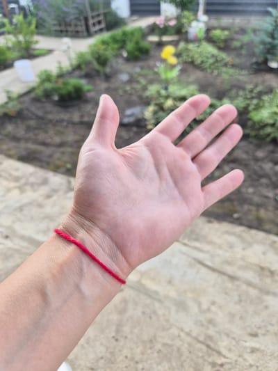 The media file displays a close-up view of a person's left hand and wrist, which is adorned with a simple red string bracelet. The hand is open, palm facing upwards, revealing the lines and texture of the skin. The wrist is positioned on a textured, light-colored surface, possibly a stone pathway or patio. In the background, a garden setting is visible, with blurred greenery, plants, and some decorative elements. The overall scene suggests an outdoor environment, likely a backyard or garden in a residential area. The lighting indicates it is daytime. There is no visible text in the image. The focus is on the hand and the red string, suggesting a potential symbolic meaning for the bracelet. The blurred background provides context without distracting from the main subject. The scene is casual and appears to be taken in a natural, everyday setting.