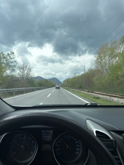 The image presents a forward view from inside a vehicle, situated on a road in Bistrice, Montenegro. In the foreground, the lower portion of a black steering wheel and dashboard are visible. The instrument cluster displays an odometer reading of 259652 km, a Trip A reading of 4724.4 km, and an ambient temperature of 16°C. Both the tachometer and speedometer needles are at 0. The fuel gauge indicates approximately a half-full tank. A black mobile phone holder is attached to an air vent on the right side of the dashboard.
Ahead, a two-lane asphalt road with white lane markings extends into the distance. A metal guardrail runs parallel to the left side of the road. On the right, a railway track parallels the road. Both sides of the road are bordered by vegetation, including trees with varying degrees of foliage. A light-colored sedan is visible in the distant lane, traveling in the same direction. Distant mountains are present on the horizon beneath a heavily overcast sky. Overhead power lines are visible against the cloudy sky on the right side of the frame.