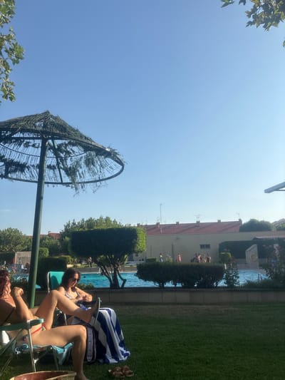 The image depicts an outdoor scene, likely a community pool or recreational area, in Villa Del Prado, Spain. In the foreground, two individuals are seated in lounge chairs on a well-maintained green lawn, enjoying a sunny day. The person on the left, with reddish-brown hair, is partially visible, wearing a dark bikini top. The person on the right, with dark hair and sunglasses, is wearing a patterned bikini top and appears to be interacting with her phone. Both are covered partially by a blue and white striped towel draped over their chairs. A large, rustic thatched umbrella stands to the left, providing shade.
In the mid-ground, a swimming pool is visible, with people scattered around its edge. Some are standing, while others appear to be entering or exiting the water. The pool area is bordered by a low concrete wall and manicured hedges. Beyond the pool, buildings with terracotta roofs are visible, characteristic of Spanish architecture. The sky is a clear, bright blue, suggesting a warm and pleasant day. The overall atmosphere is one of relaxation and leisure.