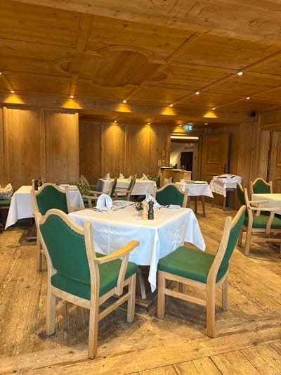 This image depicts the interior of an unoccupied traditional dining room, identified as a "Gaststube," located in Going am Wilden Kaiser, Austria. The room features a prominent alpine-style aesthetic characterized by extensive use of light-colored wood. The floor is constructed from wide wooden planks. The walls are paneled with vertical wooden boards and include recessed sections, all in a uniform light wood tone. The ceiling is coffered with carved wooden elements and features multiple recessed spotlights providing warm illumination.
Several dining tables, each draped with a white tablecloth, are arranged throughout the room. The tables are set with white folded napkins, plates, and cutlery. On the central foreground table, a set of wooden salt and pepper mills and a small decorative item (possibly a flower in a vase) are present. The chairs are constructed from light wood with dark green upholstered seats and backs, many featuring brass stud detailing along the edges.
In the background, a wooden door, partially ajar, leads into an adjacent area. Above this doorway, a green emergency exit sign is visible. The word "Gaststube" is inscribed on or above the doorway. No individuals are present in the scene, and no specific actions or interactions are occurring. The environment appears prepared for guest service.