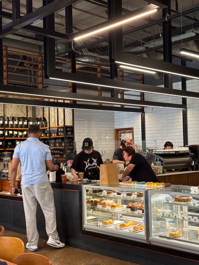 The image captures an indoor scene, likely a cafe or bakery, with a modern industrial aesthetic characterized by exposed pipes, metal beams, and warm wooden accents. The lighting is bright, with long, linear fluorescent lights illuminating the space.

Several people are visible, engaged in various activities. In the foreground, a person wearing a light blue polo shirt and grey sweatpants is facing away from the camera, holding a phone. Behind them, another individual in a black t-shirt with a white graphic design and a black baseball cap is behind a counter, seemingly interacting with a customer or preparing something. Further back, another person in a black t-shirt and a beige baseball cap is also near the counter. A glimpse of another person with dark hair can be seen behind a coffee machine.

A prominent feature of the scene is a refrigerated display case filled with various pastries and desserts, including cakes and croissants. Bags of what appear to be coffee or other products are stacked on shelves in the background.

The overall atmosphere seems casual and busy, suggesting it might be during operating hours. The environment appears clean and well-lit. There is no visible indication of the time of day or weather outside.