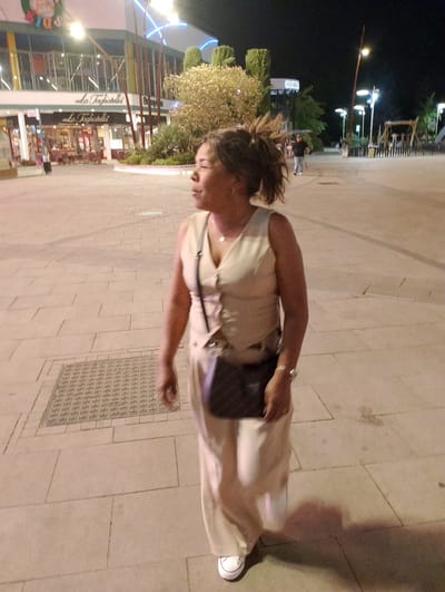 This media file shows a woman walking on a paved outdoor area at night. She is wearing a light-colored sleeveless dress and a dark crossbody bag. She also wears a watch and a ring. The woman has her hair in a bun and is looking to her right. The background shows a building with "La Tagliatella" written on it, suggesting it's a restaurant. There are also streetlights and trees visible. The setting appears to be a public space, possibly a shopping area or promenade. The time of day is clearly night due to the darkness and artificial lighting. The overall mood is neutral, with the focus on the woman and her surroundings. There is no information about the weather or the specific emotions of the subject. The location context provided states "Alcorcón, Spain," which helps to contextualize the scene.