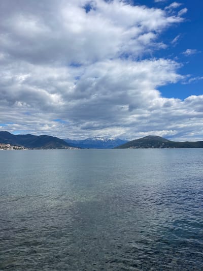 The image shows a landscape with a body of water, mountains, and sky. The water is calm with a slight ripple effect on the surface. A mountain range separates the water from the sky. The sky is blue with patches of white clouds. The location is Provodina, Montenegro. There are no people visible in the image. The scene appears to be taken during daylight hours.
