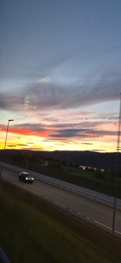 The image depicts a dramatic sunset over a highway in Biri, Norway. The sky is a vibrant canvas of colors, with streaks of pink, orange, and yellow clouds against a deepening blue. A silhouette of hills forms the horizon, with hints of trees and a body of water reflecting the fading light.

In the foreground, a highway stretches into the distance, with a single car driving on it, its headlights illuminating the road. The road has clear lane markings and a metal barrier separating it from the grassy shoulder. A streetlamp is visible on the left side of the image.

The overall mood is serene and awe-inspiring, capturing the beauty of nature at dusk. The location is identifiable as Biri, Norway. The text "Klein" is visible vertically on a sign in the sky, likely a billboard. The time of day is clearly sunset.
