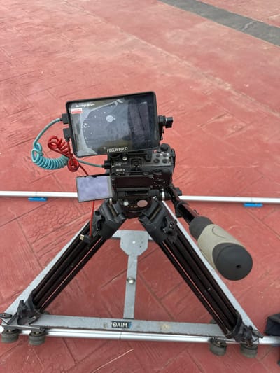 The image shows a Sony FX30 camera with a FEELWORLD monitor mounted on a tripod slider. The camera and monitor are set up on a red, brick-like paved surface. Two coiled cables, one blue and one red, are connected to the monitor. The slider has silver metal rails and black legs, with wheels visible at the bottom. The background is a continuation of the red paved surface. No people are visible in the image. The scene appears to be outdoors, possibly in a public area.
