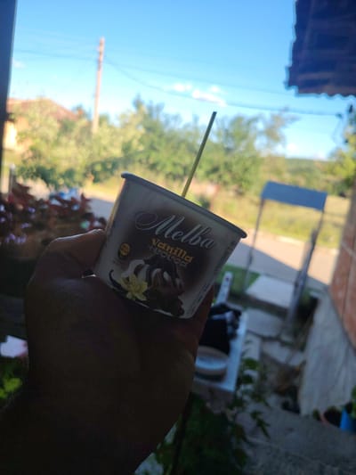 A hand holds a Melba vanilla and cocoa ice cream cup with a straw inserted. The cup is partially obscured by the hand and the background is blurred, showing a building with a brick facade and a blue roofed structure, trees, and a utility pole against a bright blue sky. The setting appears to be Borintsi, Bulgaria, possibly a residential area due to the presence of a house and a yard. The lighting suggests it is daytime, with the sun casting shadows. The overall impression is of a casual moment outdoors, perhaps on a porch or patio.