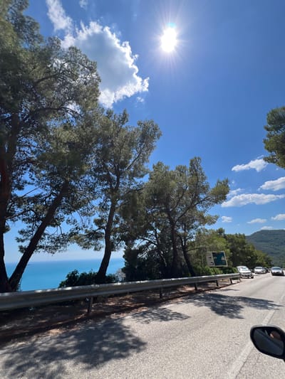 The image shows a scenic view of a road in Mattinata, Italy, with the sea visible in the background. The sun is shining brightly in a clear blue sky with some white clouds. On the left side of the road, there are several tall pine trees with dense foliage. A metal guardrail separates the road from the cliff edge. Cars are driving on the road, and there is a signpost with some symbols. The road appears to be paved with asphalt and shows some shadows from the trees. The sea is a vibrant blue, and the coastline is lined with greenery. The overall scene is one of a beautiful coastal drive on a sunny day.