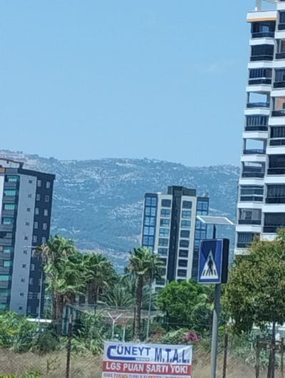 This image depicts an urban landscape with multiple high-rise buildings, trees, and a street sign. In the foreground, a banner with Turkish text reads "CÜNEYT M.T.A.L. LGS PUAN ŞARTI YOK!". To the right, a blue and white pedestrian crossing sign is mounted on a pole. Several palm trees and other green foliage are visible. In the background, a range of rocky, treeless hills is under a clear blue sky. The buildings are modern, with dark-framed windows and balconies. The overall scene suggests a sunny day in a city, likely Toroslar, Turkey, given the presence of Turkish text. There are no people clearly visible in the image.