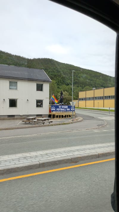 The image shows a street scene in Mosjøen, Norway. On the left is a white, two-story building with several windows. In front of the building, there is a wooden deck with picnic tables and umbrellas. A rainbow flag is flying on the deck. A sign on the deck reads "VI VISER FOTBALL VM". To the right of the building is a yellow, industrial-looking building with a row of windows. The street in front of the buildings is paved and there is a pedestrian walkway with a yellow line. A forested hill rises in the background. The sky is overcast.
