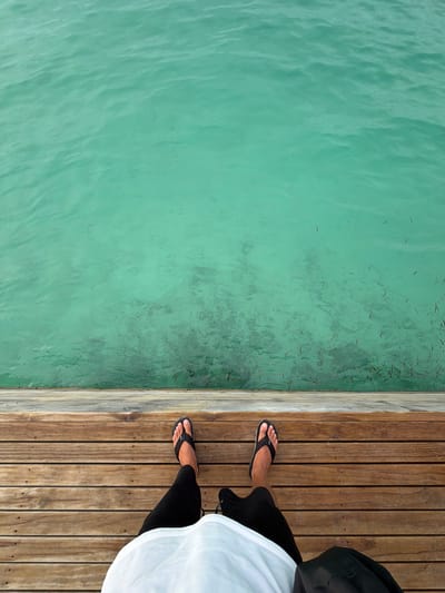 View of turquoise water and a wooden dock on Guraidhoo, Maldives. A person wearing black sandals, black pants, and a white shirt stands on the dock.