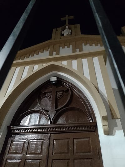 The image captures the front facade of a church at night, likely in Quilmes, Argentina. The scene is partially obstructed by what appear to be metal fence bars in the foreground, blurring the clarity of the full view. The church entrance has a large, dark wooden double door with an arched top featuring a prominent cross design. Above the arch is a modern security light fixture. The building's facade is painted in alternating bands of beige and white, with a decorative, crenellated roofline. Perched on the roof above the entrance is a small statue, possibly of Jesus or another religious figure, with a Christian cross mounted above it. The darkness of the sky suggests a nighttime setting.